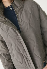 Close-up van een persoon in een grijze gewatteerde jas over een marineblauwe ritstrui, waarbij de voorkant van de rits en de knoopdetails zichtbaar zijn.