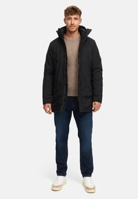 Giacca nera imbottita con colletto alto, abbinata a un maglione marrone chiaro, jeans blu e sneakers bianche. Sfondo semplice.
