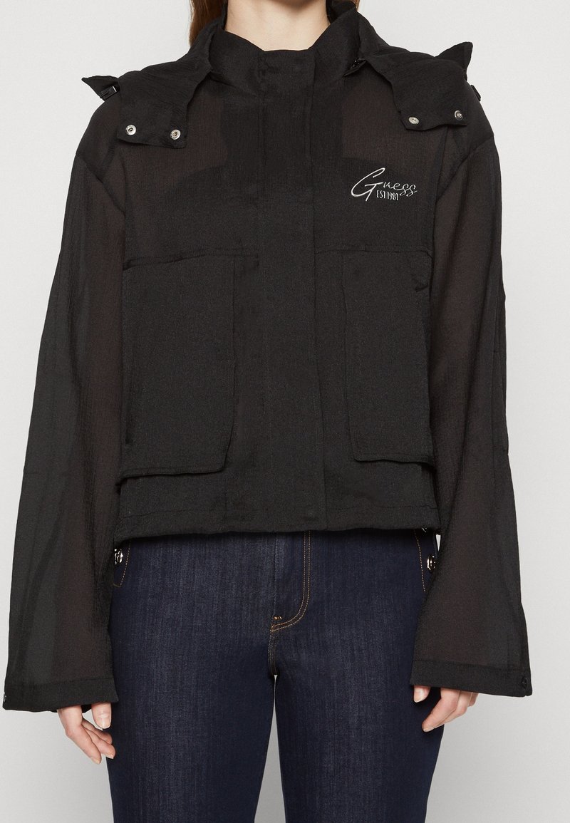 Personne portant une veste Guess noire avec de grandes poches avant et un logo sur la poitrine, associée à un jean bleu foncé, debout devant un fond uni.