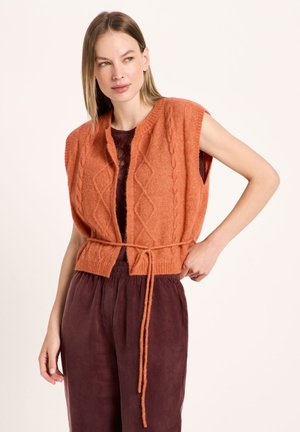 Woman wearing a sleeveless orange knit vest with a tie belt over a burgundy top and matching burgundy pants, looking to the side.