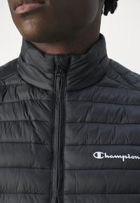 Detalní záběr na osobu ve černé prošívané bundě zapnuté až k krku s bílým logem Champion na hrudi.