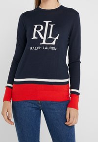 Frau, die einen marineblauen Ralph Lauren Pullover mit weißen und roten Streifen und Logo trägt, kombiniert mit blauen Jeans, steht vor einem einfarbigen Hintergrund.