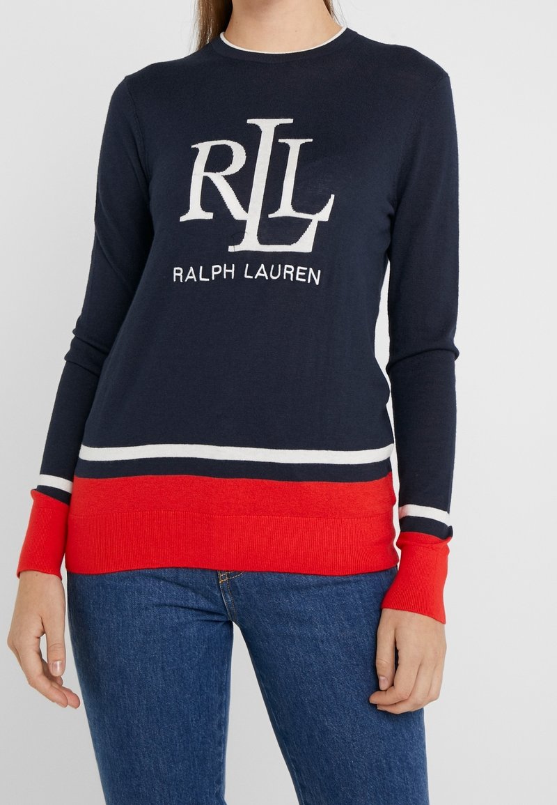 Frau, die einen marineblauen Ralph Lauren Pullover mit weißen und roten Streifen und Logo trägt, kombiniert mit blauen Jeans, steht vor einem einfarbigen Hintergrund.