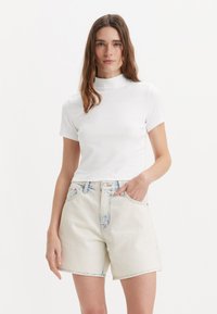 Un haut à col montant blanc à manches courtes associé à un short en jean taille haute de couleur claire. Le tissu semble lisse avec un minimum de déchirures.