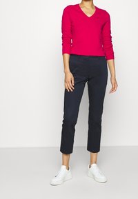 Maglione rosa a V con maniche lunghe abbinato a pantaloni cropped blu scuro e sneakers bianche. Design semplice senza motivi visibili.