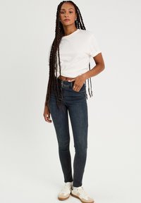 Eng anliegende dunkelblaue Jeans kombiniert mit einem weißen Kurzarm-T-Shirt. Sie trägt cremefarbene und beiger Sneakers und steht vor einem schlichten Hintergrund.