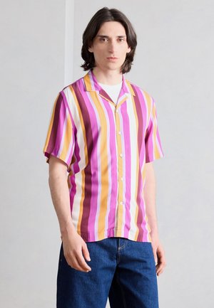 Άνδρας με μαλλιά μήκους ώμων, που φοράει κοντομάνικο πουκάμισο με κάθετες ριγέ σε ροζ, κίτρινο, λευκό και μωβ χρώμα και μπλε τζιν.