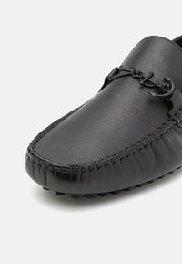 Mocassin en cuir noir avec un dessus texturé, pointe moc en couture et un détail métallique en forme d'ancre décoratif sur fond blanc.