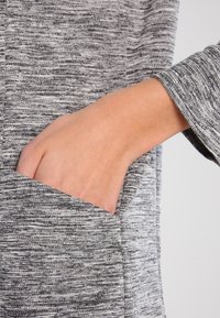 Šedý pletený top s kapsou, detail ruky spočívající v kapse. Textura je měkká s melírovaným vzorem.