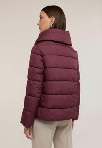 Žena s rovnými hnědými vlasy, která nosí stříbrné kruhové náušnice a vínově červenou prošívanou péřovou bundu, béžové kalhoty, otočená do boku.