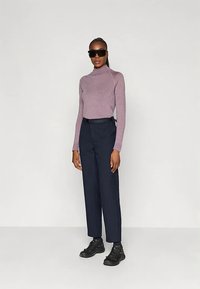 Moteris stovi, vilkėdama juodus akinius nuo saulės, mauve spalvos ilgomis rankovėmis turtleneku, juodą diržą, tamsiai mėlynus kelnes ir juodus batus, prieš baltą foną.