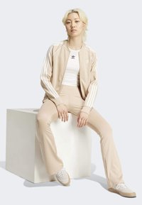 Chándal deportivo beige con rayas blancas. Pantalones largos de corte ajustado y chaqueta a juego. Modelo sentado en un pedestal blanco, llevando zapatillas blancas.