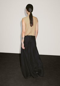 Beige ärmelloses Top, kombiniert mit einem langen, fließenden schwarzen Rock. Der Rock hat einen sichtbaren Reißverschluss auf der Rückseite und strukturierte Stoff.