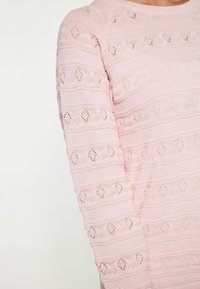 Maglione lavorato a maglia rosa chiaro con motivi testurizzati e design a rete, caratterizzato da righe orizzontali e un collo rotondo.