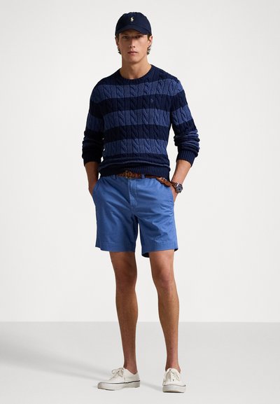Marineblå og blå stribet kabelstrikket sweater, lyseblå shorts, brun bælte, hvide sneakers og marineblå kasket med logo. Enkel, afslappet påklædning.