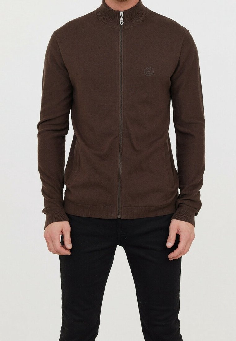 Homme portant un pull marron à manches longues zippé avec un col montant et un pantalon noir sur un fond uni.