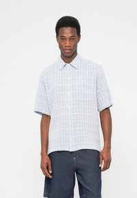 Άνδρας που φοράει ανοιχτό μπλε κοντομάνικο πουκάμισο με καρό μοτίβο και σκούρα μπλε σορτς από τζιν, στέκεται μπροστά σε λευκό φόντο.