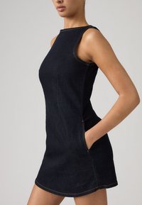 Γυναίκα που φοράει ένα αμάνικο σκούρο φόρεμα από τζιν με αντίθετες ραφές, με το δεξί της χέρι στην πλαϊνή τσέπη, μπροστά από απλό φόντο.