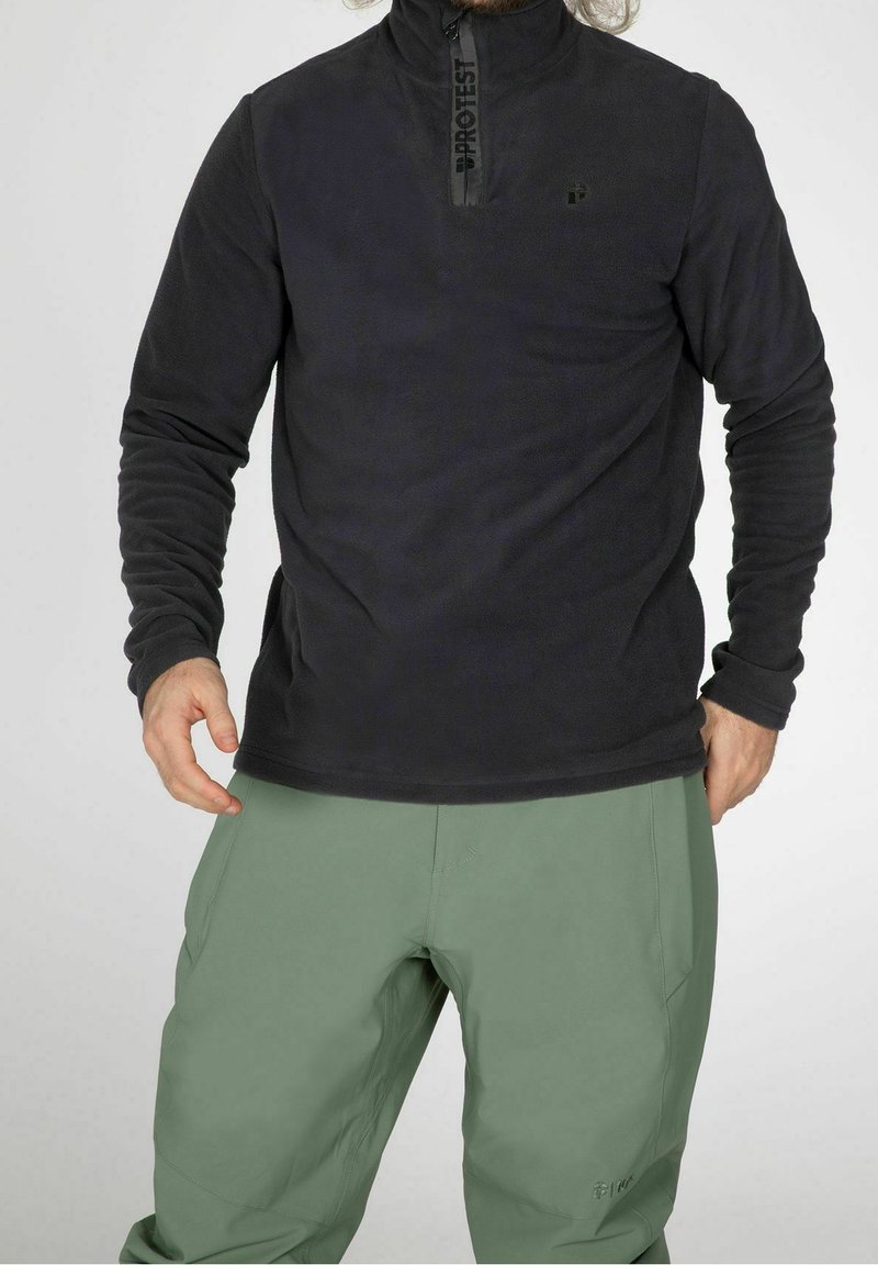 Pull en polaire noir avec col zippé, présentant un logo sur la fermeture éclair. Associé à un pantalon vert clair, offrant une coupe décontractée et une texture.