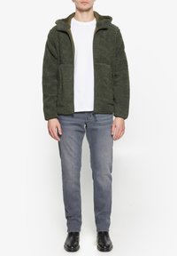 Giacca in pile verde con zip e cappuccio, dotata di due tasche frontali, abbinata a jeans grigi e stivali neri. Design semplice e testurizzato.
