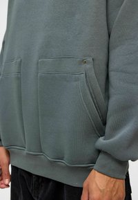 Grijze sweatshirt met twee grote voorzakken, geribde manchetten en een gladde textuur, voorzien van accentstiksels en een metalen drukknoopdetail.