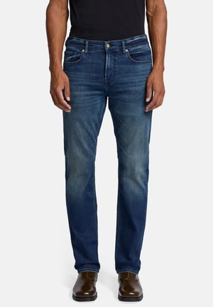 Jeans in denim blu scuro con una vestibilità a gamba dritta, leggere sfumature sulle cosce, cinque tasche e chiusura con bottone in metallo. Indossati con stivaletti marroni fino alla caviglia.