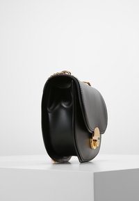 Musta nahkainen olkalaukku, jossa on läppäkansi, kultaiset metalliosat ja ketjuhihna, sileällä tekstuurilla ja kaarevalla muodolla.