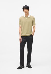 Pikétröja i ljus olivgrön med vit krage och dragkedja, kombinerad med svarta byxor och svarta skor. Enkel, figurnära design.