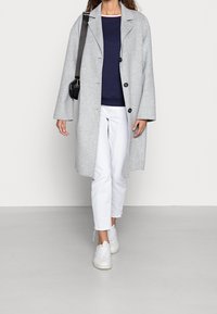 Šedý vlněný kabát s velkými knoflíky, nošený přes modrý svetr, v kombinaci s bílými zkrácenými kalhotami a bílými teniskami. Jednoduchý design, uvolněný střih.