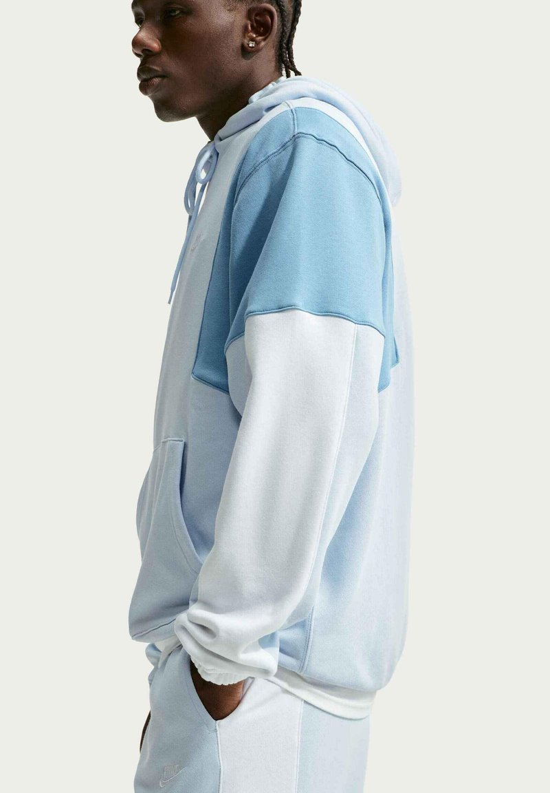 Persoon in profiel draagt een lichtblauwe en witte colorblock-hoodie met bijpassende joggingbroek, met de handen in de zakken tegen een effen achtergrond.
