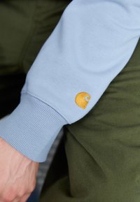 Lyseblå sweatshirtærme med ribbet manchet og et lille gult logo. Baggrunden består af mørkegrønt stof, hvilket tilføjer farvekontrast.