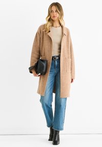 Oversized brown wool coat with notch lapels, two front pockets; paired with a beige top, blue jeans, and black ankle boots.