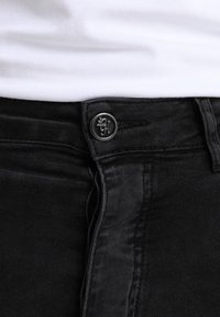 Jeans de mezclilla negra con una textura suave, que presentan un botón de metal con un logotipo en relieve y un diseño de cremallera abierta.