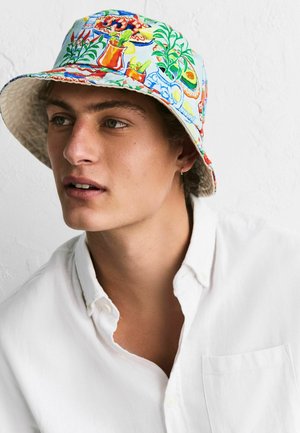 Nuori mies, jolla on lyhyet kiharat hiukset, valkoinen nappipaita ja värikäs kalastajanhattu, jossa on kasvi- ja ruokakuvituksia.