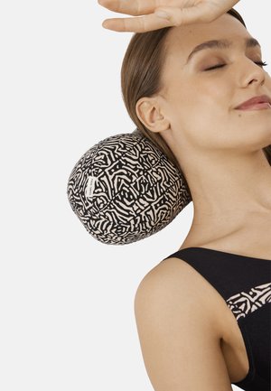 Mujer con los ojos cerrados descansando el cuello sobre una almohada redonda con patrón en blanco y negro, vestida con una blusa negra que tiene un patrón a juego.