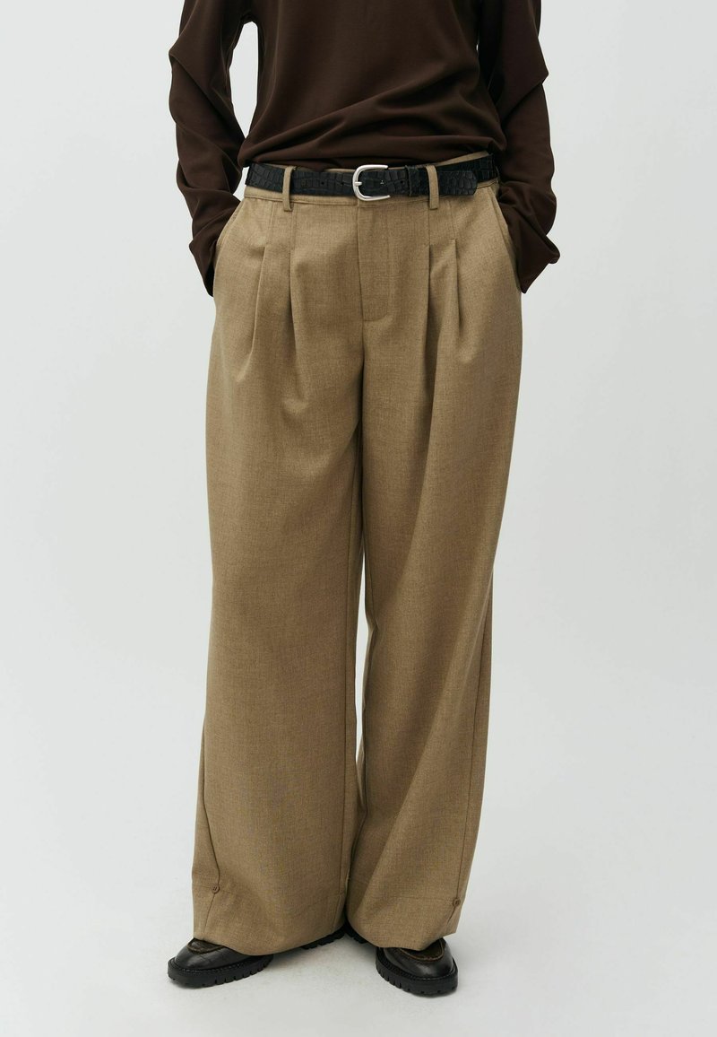 Personne portant un pantalon beige à jambes larges, une ceinture noire texturée, un haut à manches longues marron foncé et des chaussures noires, debout les mains dans les poches.