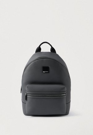 Mochila cinza em pele sintética com forma arredondada, bolso frontal com fecho, logótipo em preto como detalhe e alça superior para transporte. Textura suave em geral.