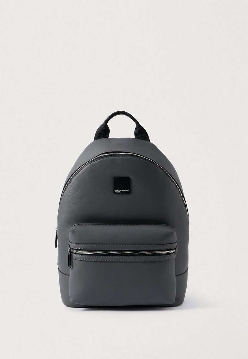 Pelēka mākslīgās ādas mugursoma ar noapaļotu formu, priekšējo rāvējslēdzēja kabatu, melnu akcentu logotipu un augšējo rokturi pārvadāšanai. Virsma ir gluda.