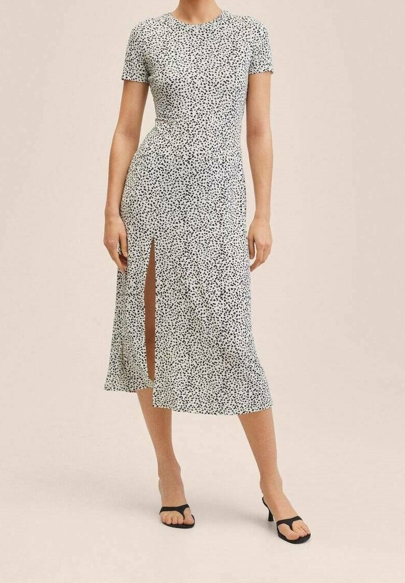 Femme portant une robe midi blanche à pois noirs, manches courtes, fente latérale, et sandales à talons noires ouvertes à l’avant, debout sur un fond beige.