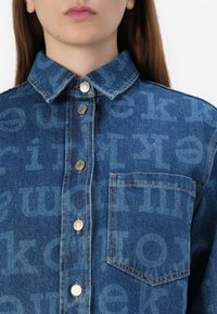 Et nærbilde av en person som har på seg en denimskjorte med knappelukking og brystlomme, med et svakt gjentatt tekstmønster.