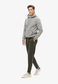 Grijze hoodie met een voorzak en een trekkoord, gecombineerd met donkergroene joggingbroek. Model draagt lichtgrijze sneakers met een textuur.