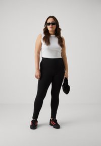 Black high-waisted leggings, fitted design, paired with a white sleeveless top. Worn with black sneakers featuring red accents, holding a black accessory.
