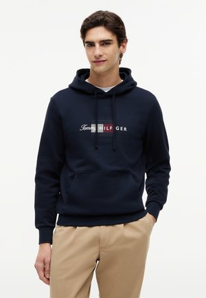 Junger Mann trägt einen dunkelblauen Hoodie mit einer Vorderseite-Tasche und Logotext, kombiniert mit beigen Hosen, und steht vor einem schlichten Hintergrund.