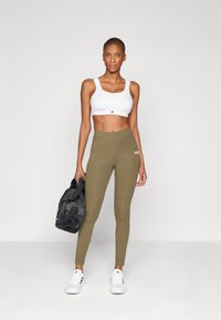Vit sportbh, olivgröna leggings och vita sneakers. Modellen håller en svart duffelväska. Släta, tighta tyger med logotyper.