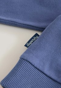 Sudadera azul oscura con tejido texturizado, puños acanalados y una pequeña etiqueta blanca con el logo que dice "Superdry" cosida en la manga.