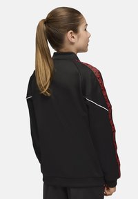 Chaqueta atlética negra con diseños geométricos rojos en los hombros, ribetes blancos y un cuello alto. Tela suave, corte relajado.
