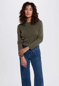 Olijfgroene longsleeve top met een geschulpte halslijn en versierde manchetten, gecombineerd met hoge blauwe denimspijkerbroeken. Hete zachte textuur.