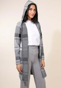 Cardigan à capuche gris avec un motif à carreaux, présentant une coupe ample et des manches longues. Fabriqué en tissu doux, associé à un pantalon gris.