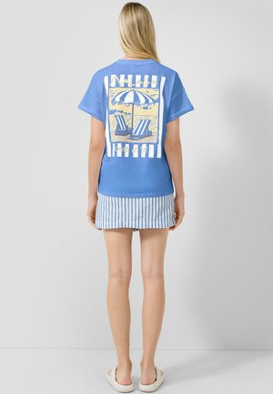 Kobieta stojąca tyłem, ubrana w niebieski T-shirt z motywem plaży i napisem „Malaga Costa del Sol”, spódnicę w paski i białe buty na wsuwanie.