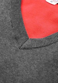 Grauer Pullover mit V-Ausschnitt, der einen strukturierten Rippkragen und ein rotes Innenfutter hat. Grösse-Schild sichtbar, Grösse M.